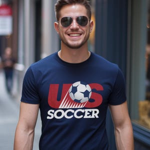 May include: A navy blue short-sleeved t-shirt featuring a graphic design on the chest. The design includes the red letters US with a white and black soccer ball positioned over the letter S, and the word SOCCER printed in white below the letters.