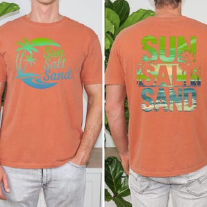 K&ouml;nnte beinhalten: Gebrannt orangefarbenes T-Shirt mit dem Schriftzug "Sun Salt Sand" in einem tropischen Design auf Vorder- und R&uuml;ckseite. Das Design umfasst Palmen und Meereswellen in Gr&uuml;n, Blau und Wei&szlig;.