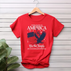 May include: A red t-shirt with a patriotic design. The shirt features the text "UNITED STATES OF AMERICA" above an illustration of an American flag, an eagle, and the Statue of Liberty. The text "We the People" and "250-YEAR SEMI-QUINCENTENNIAL" are also included.