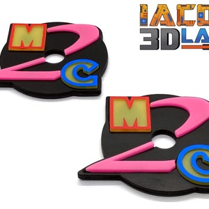 May include: Two black plastic keychains with a pink and yellow design. The keychains feature the letters "M" and "C" with a pink curved line. The text "IACOM 3D LABS EST. 2021" is visible in the background.
