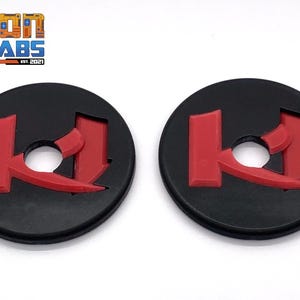 Puede incluir: Dos discos redondos de plástico negro con una letra K estilizada roja en el centro. Los discos tienen un agujero en el centro.