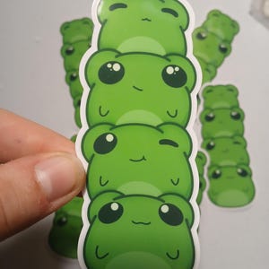 Op de afbeelding: Een stapel groene kikkerstickers met witte randen. De stickers tonen cartoon kikkergezichten met grote ogen en lachende monden. Op de achtergrond zijn nog een aantal kikkerstickers te zien.
