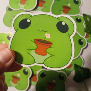 May include: A collection of green frog stickers. Each sticker features a cartoon frog holding a small orange pot with a flower. The frog has large black eyes and a smiling expression. The stickers are on a white background.