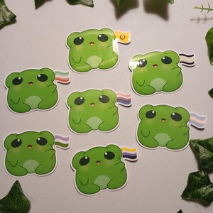 May include: Seven green frog stickers, each with black eyes and a small pink mouth, holding different pride flags. The stickers are on a white surface, decorated with green leaves. The frog stickers are a fun and colourful design.