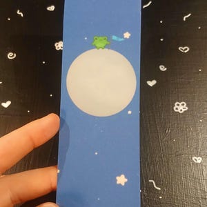 Peut inclure: Un marque-page bleu avec une lune blanche et une grenouille verte tenant une étoile bleue. L'arrière-plan est bleu foncé avec des étoiles blanches.