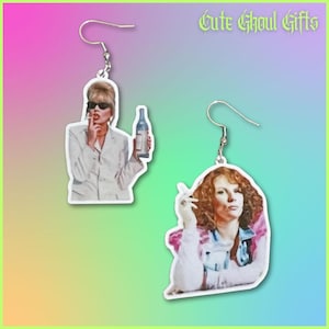 May include: Dangle earrings with images of two women. One earring depicts a woman in a white suit holding a bottle and smoking. The other earring shows a woman with red hair smoking. The text "Cute Ghoul Gifts" is in the top right corner of the image.