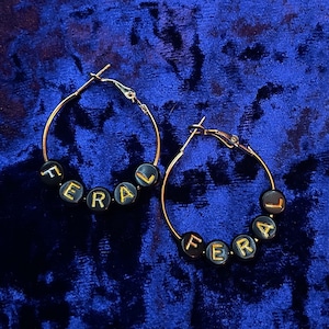 May include: A pair of gold hoop earrings with black beads spelling out the word "FERAL" in gold lettering.