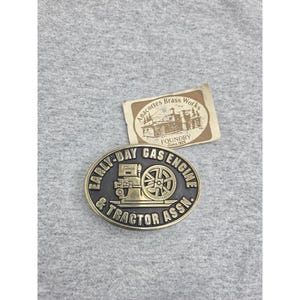 May include: An oval brass belt buckle featuring a raised relief of a stationary engine and the text EARLY-DAY GAS ENGINE & TRACTOR ASSN. It is accompanied by a rectangular paper tag labelled Anacortes Brass Works Foundry Since 1926.