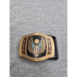 May include: A rectangular belt buckle with a brushed gold-tone finish featuring a central black panel. The design includes a winged scarab motif with turquoise inlay, a circular sun disc, and side panels with red accents and stylized floral patterns.