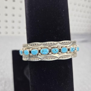 Puede incluir: Un brazalete de plata con una fila central de piedras turquesa ovaladas. La banda de metal está decorada con patrones geométricos estampados a lo largo de los bordes superior e inferior, con pequeñas cuentas de plata entre cada piedra turquesa.
