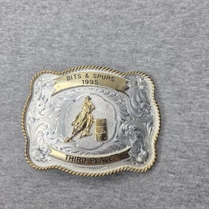 May include: A rectangular silver-tone belt buckle with a gold-tone rope border. It features an engraved floral pattern, a gold-tone figure of a rider on a horse by a barrel, and two gold-tone banners reading BITS & SPURS 1995 and THIRD PLACE.