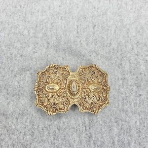 May include: A gold-toned metal belt buckle featuring an intricate filigree pattern with scrolling lines and textured details. The rectangular shape has scalloped edges and a central oval medallion flanked by two smaller oval accents on each side.