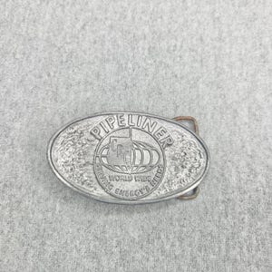 Peut inclure: Une boucle de ceinture ovale en métal argenté avec une bordure texturée et un emblème central. Le texte PIPELINER est arqué en haut, avec CRC au centre d'un graphique de globe et les mots SERVING WORLD WIDE ENERGY'S NEEDS inscrits en dessous.