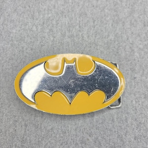 Puede incluir: Una hebilla de cinturón ovalada con el logotipo de Batman. El diseño consta de un borde de esmalte amarillo brillante y una silueta central de murciélago en metal plateado. Una hebilla metálica rectangular está unida al lado derecho.