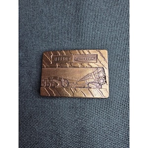 May include: A rectangular bronze-toned belt buckle featuring an embossed design of heavy machinery. The text JEFFREY DRESSER is clearly visible at the top. The background of the buckle has a textured pattern resembling tire treads.