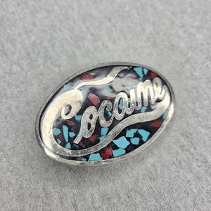 May include: An oval-shaped metal pin featuring the word "Cocaine" in raised silver-toned script lettering. The background is filled with a mosaic of small, crushed turquoise and red stone fragments set within a dark, polished resin base.