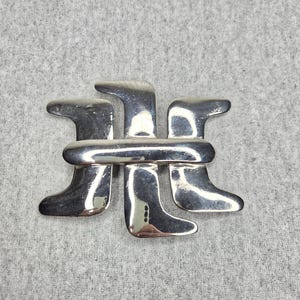Puede incluir: Una hebilla de cinturón de metal plateado pulido con un diseño abstracto y estilizado. La hebilla consta de una barra horizontal que cruza tres formas verticales curvas similares a botas que apuntan en direcciones alternas, creando un accesorio metálico simétrico, geométrico y reflectante.