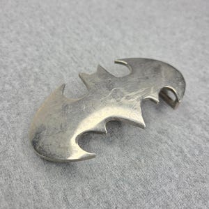 Op de afbeelding: Een zilverkleurige metalen riemgesp met een gestileerd, symmetrisch vleermuisachtig silhouet met puntige vleugels en een centraal lichaam. Het oppervlak heeft een gladde, reflecterende afwerking met zichtbare lichte krassen.