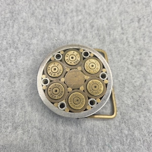 May include: A circular belt buckle featuring a silver-toned metal frame containing five brass-colored 44 REM MAG cartridge casings arranged around a central circular brass disc, with small silver metal rings interspersed between the casings.