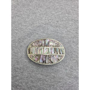 Pode incluir: Uma fivela de cinto oval com uma moldura metálica e incrustações de segmentos de concha de abalone iridescente. O design inclui uma barra central horizontal que divide as peças de concha, exibindo tons perolados naturais.