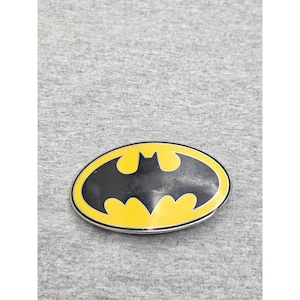 Puede incluir: Una hebilla de cinturón de forma ovalada que presenta una silueta del logotipo de Batman en negro centrada sobre un fondo amarillo brillante, rodeada por un borde fino de metal plateado. La superficie muestra signos de desgaste.