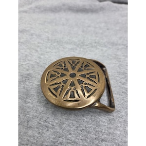 May include: A circular brass belt buckle featuring an intricate openwork design of radiating spokes and geometric cutouts. The metal has a warm, aged patina with visible dark recessed areas between the raised, polished brass patterns.