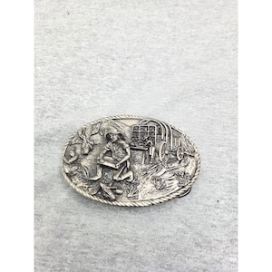 May include: An oval-shaped silver-toned metal belt buckle featuring a raised relief scene of a person panning for gold in a stream near a covered wagon. The border of the buckle has a twisted rope design, and the entire surface has a metallic finish.
