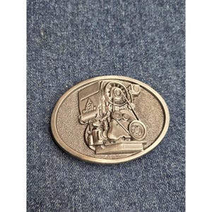 May include: Oval silver belt buckle with a detailed engine relief and the text "KHD DEUTZ". The buckle has a textured surface and a classic design, suitable for a belt. The buckle is made of metal.