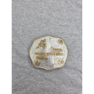 Op de afbeelding: Een zilver- en goudkleurige riemgesp met een gegraveerd bloemmotief, een figuur van een ruiter op een paard en de tekst FINALS HELL ON WHEELS RODEO CHAMPION 1998. De gesp heeft een touwachtige rand en twee kleine bloemaccenten.