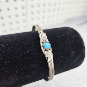 May include: A silver cuff bracelet featuring a central round turquoise stone set in a serrated bezel. The metal band is decorated with two symmetrical leaf-like engravings flanking the stone, creating a detailed and polished metallic finish.