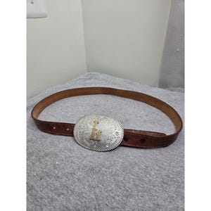 May include: A brown leather belt with a textured lizard-skin pattern and a large oval silver-tone metal buckle. The buckle features an intricate floral engraving and a prominent gold-tone letter H centered on the front surface of the buckle.