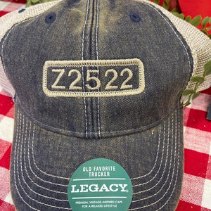 May include: A blue and brown trucker hat with a mesh back. The hat has a tan patch with the text "Z2522" embroidered on it. The hat also has a green circular patch with the text "OLD FAVORITE TRUCKER LEGACY" and the text "PREMIUM, VINTAGE-INSPIRED CAPS FOR A RELAXED LIFESTYLE #LIVEYOURLEGACY".