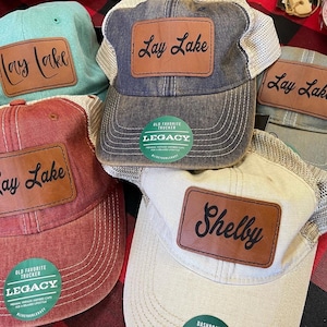 May include: Four trucker hats with brown leather patches. The patches have the text "Lay Lake" and "Shelby" in black script. The hats are in various colors, including blue, red, green, and beige. The hats have a mesh back and a logo that says "Old Favorite Trucker Legacy".