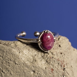 May include: A silver ring with a pink and purple oval gemstone set in a silver bezel. The ring is sitting on a rough, grey surface.