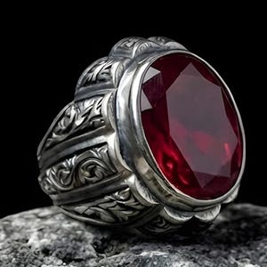 Puede incluir: Anillo de plata con una gema roja oscura ovalada y facetada. El anillo tiene un diseño ornamentado y grabado alrededor de la banda y el engaste. Se muestra sobre una piedra gris texturizada sobre un fondo negro.