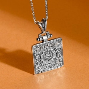 May include: A silver pendant necklace with a square, floral-engraved pendant. The pendant features a detailed rose design in the center, surrounded by ornate patterns. The necklace hangs on a delicate silver chain, creating an elegant accessory.