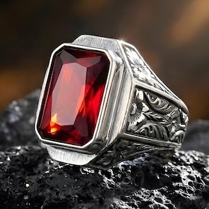 Peut inclure: Une bague en argent ornée d'une grande pierre rouge rectangulaire. La bague présente des détails floraux et des volutes complexes sur le bandeau. La pierre est sertie dans une lunette en argent, posée sur des roches sombres et texturées.