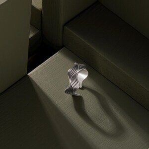 May include: A silver ring with a wavy, textured design. The ring is on a green surface.