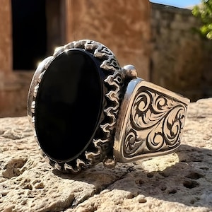 Pode incluir: Um anel de prata com uma pedra preta oval. O anel tem um design ornamentado ao redor da pedra e na banda. O anel está sobre uma superfície texturizada marrom clara.