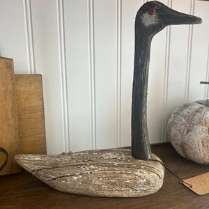 May include: A wooden carved goose figurine with a black and white painted head and a weathered brown body. The goose is standing on a wooden shelf.