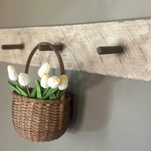 May include: A rustic wooden wall-mounted coat rack with three dark brown pegs. A woven basket filled with white and yellow tulips hangs from one of the pegs. The rack has a distressed, whitewashed finish.