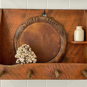 May include: A rustic, wooden wall shelf with a distressed orange finish. It features a round, carved wooden plate with the words "Spare Not" and a small shelf holding a white ceramic jar. The shelf has three wooden pegs and a decorative floral arrangement.