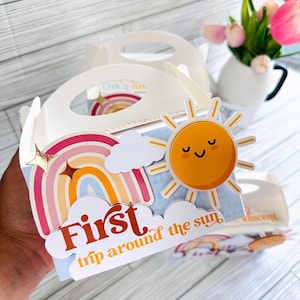 May include: White gift box with a handle, decorated with a rainbow, clouds, and a smiling sun. The text "First trip around the sun" is displayed on the front. The box is designed for a child's birthday.