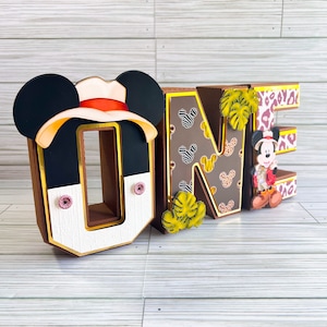 May include: A decorative "ONE" sign with a Mickey Mouse theme. The "O" features Mickey Mouse ears and a safari hat. The "N" has a zebra print pattern, and the "E" has a leopard print pattern. A Mickey Mouse figure is on the right side.