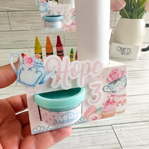 May include: A white cardboard party favor box with a pink and blue teacup design. The box has a clear plastic container with a small container of Play-Doh inside. The box also has a white paper roll and a set of crayons. The box is decorated with the name "Hope" and the number "3".