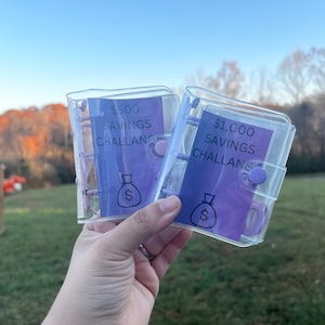 May include: Two clear plastic savings challenge binders. Each binder has a purple insert with text that reads "$500 Savings Challenge" and "$1,000 Savings Challenge". A money bag graphic is also visible.