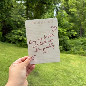 May include: A library card design with the text "Buy me books and tell me I'm pretty" in burgundy script. The card has a vintage aesthetic with lines for title, author, and due date, and includes heart doodles.