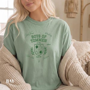 May include: A sage green short-sleeve t-shirt featuring a dark green graphic print. The design includes the text "BOYS OF TOMMEN" above a shield emblem containing shamrocks and footballs, surrounded by various handwritten-style signatures.