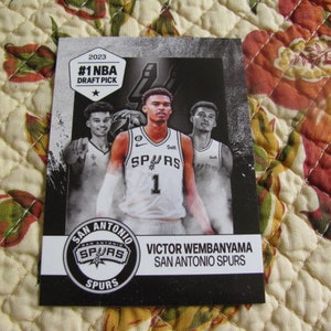 May include: A black and white basketball card featuring Victor Wembanyama of the San Antonio Spurs. The card includes the text "#1 NBA Draft Pick" and the team logo. Three players are visible in the image.