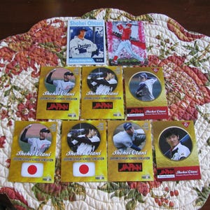 Puede incluir: Una colección de cromos de béisbol con Shohei Ohtani. Las tarjetas muestran imágenes de Ohtani en acción, con el texto "Shohei Ohtani Japanese High School Sensation" y la bandera japonesa. Las tarjetas están sobre un mantel individual con estampado floral.